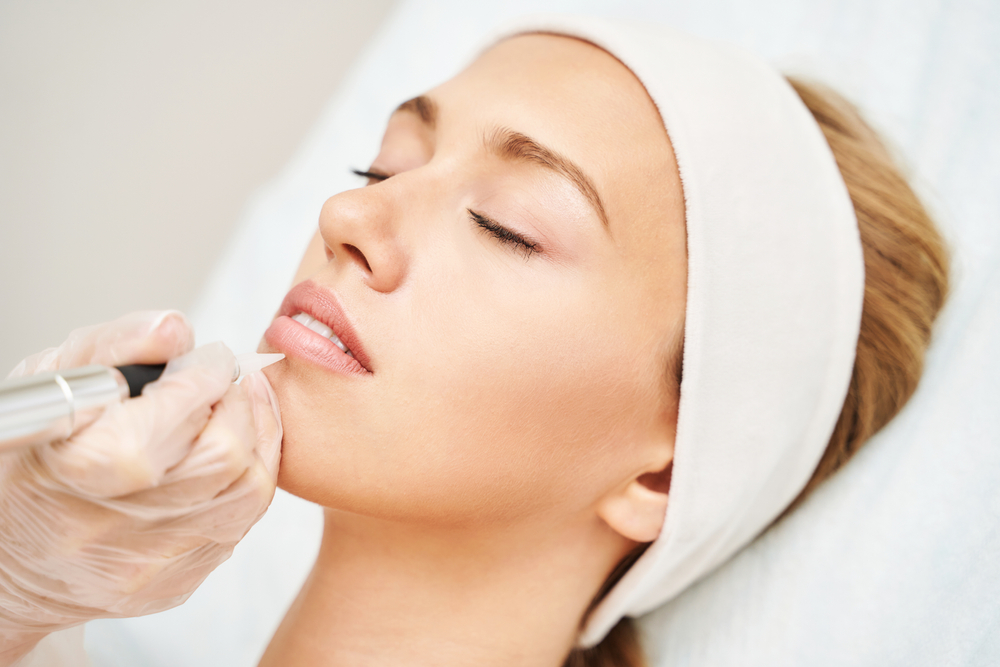 Young woman receiving lip micropigmentation at a beauty spa using a professional dermatology device for cosmetic tattooing and mouth enhancement.