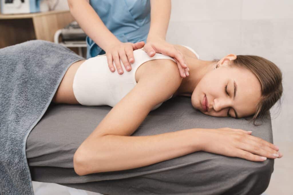 Female massage therapist performing a therapeutic back and shoulder massage on a young woman lying on a massage table, emphasizing relaxation and spa treatment benefits