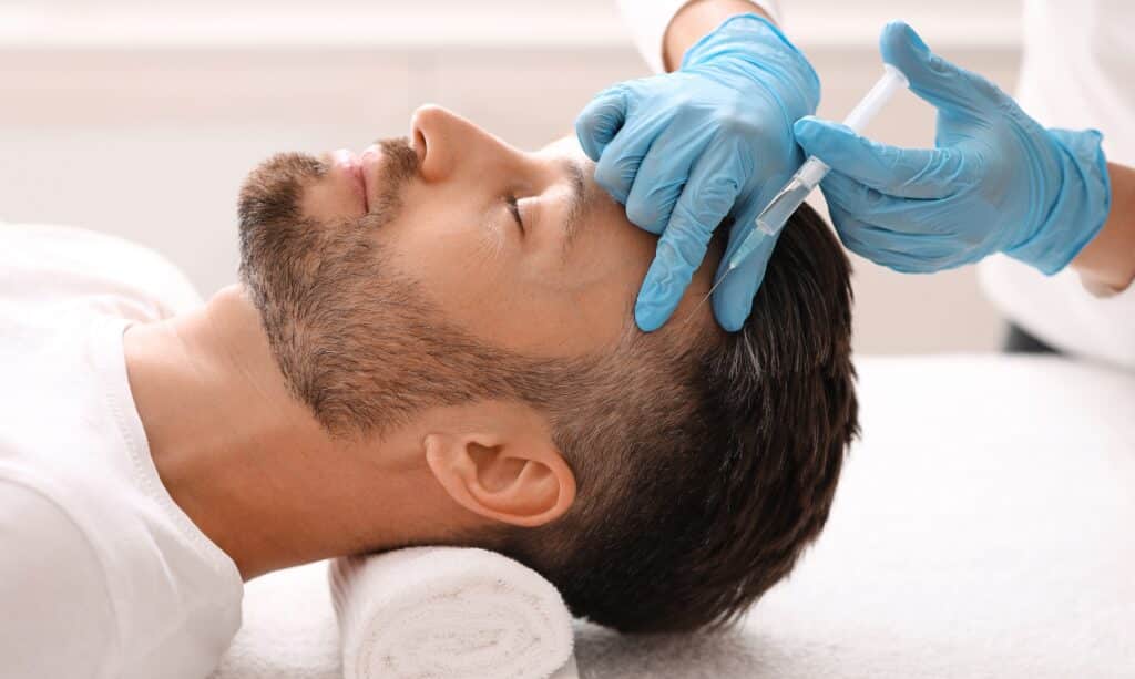 Side view of middle-aged man receiving hair treatment with scalp injection during mesotherapy session at beauty clinic, therapist’s gloved hands visible