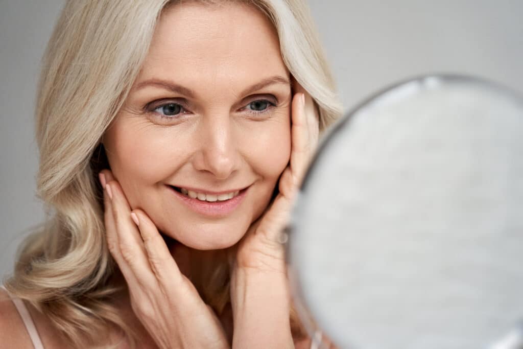 A happy middle-aged woman in her 50s touches her face while looking in the mirror, enjoying youthful skin after Thermage and injectables